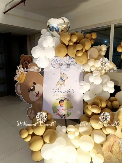 A close-up of the welcome sign for Pransh's first birthday party. A crowned teddy bear prop and a cascade of balloons create a sweet and inviting display.
