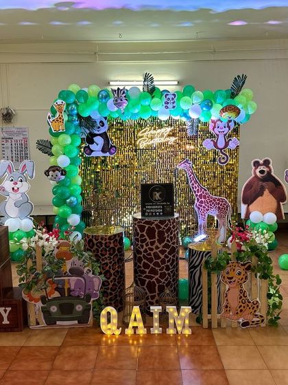 A fun jungle party setup with a golden shimmer wall and a green balloon arch. The stage is filled with character cutouts including an elephant, giraffe, and monkey, creating a playful environment.