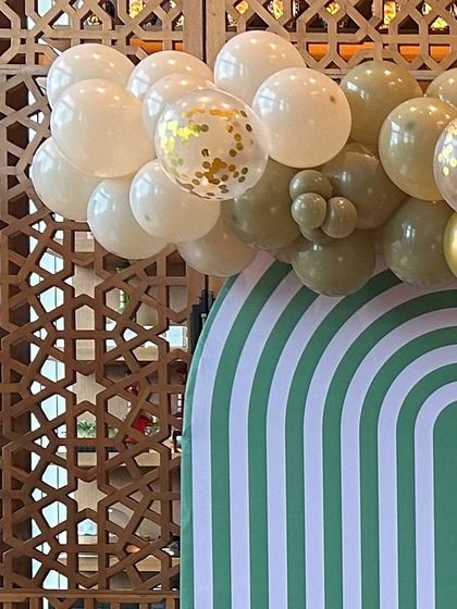 A modern and stylish backdrop with a green and white arch pattern. The setup is complemented by a balloon garland in olive green, white, and gold confetti.