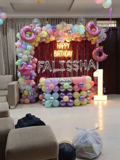 A sweet and colorful donut-themed first birthday. The backdrop is a pink tinsel curtain with a balloon arch featuring pastel colors and large donut-shaped foil balloons.