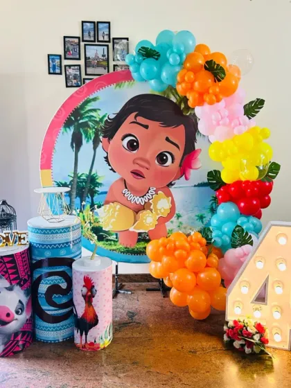 A tropical Moana-themed decoration for a 4th birthday celebration. The setup is built around a circular backdrop of Moana, with a colorful balloon garland, character-themed pedestals, and a large light-up number '4'.