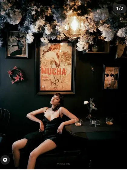 A dark, atmospheric shot in a cafe setting. The makeup is glamorous and sultry, designed to fit the moody, cinematic lighting of the scene.
