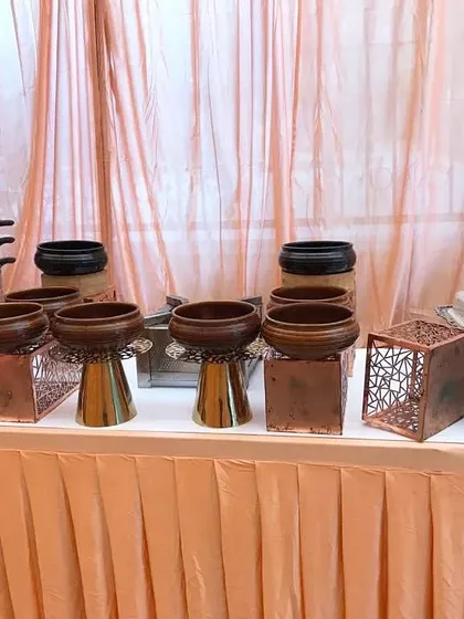 A catering station featuring a variety of rustic and modern serveware, including wooden bowls on copper stands and a multi-tiered steamer, set against a peach-colored drape.