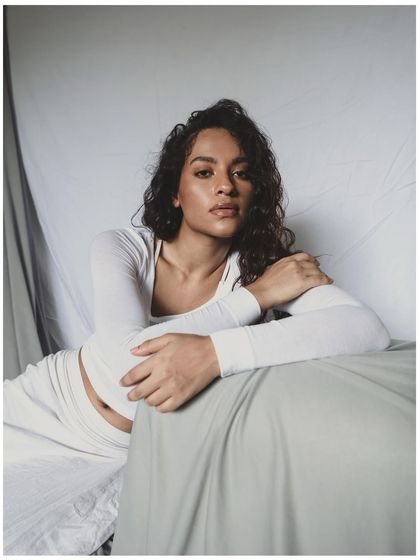 Another shot from the minimal all-white series. The model's direct gaze creates a strong connection with the viewer, contrasting with the soft, dreamy quality of the overall setup.