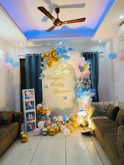 A personalized 'Oh Baby' shower decoration set up in a client's living room. The design features a custom backdrop with the mother's name, a balloon arch in pink, blue, and gold, and butterfly details.