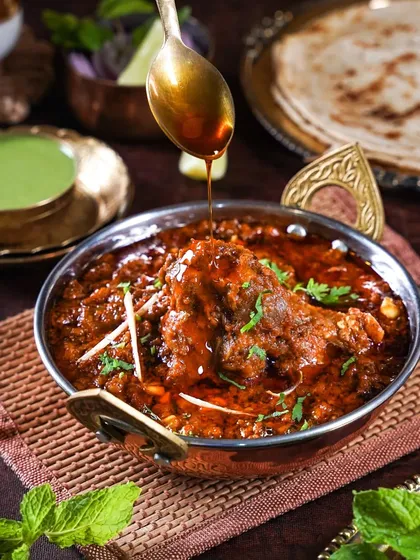 An action shot of ghee being drizzled into a bowl of spicy mutton curry. The close-up on the glistening sauce and tender meat is designed to make you crave a bite.