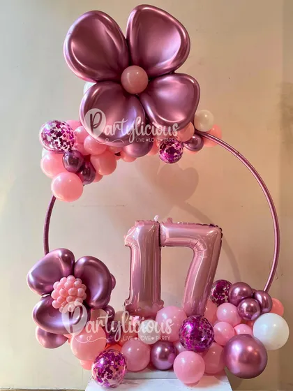Pretty in pink for a 17th birthday. This arrangement uses a metal hoop as a frame, decorated with pink and magenta balloons, floral accents, and a number display.