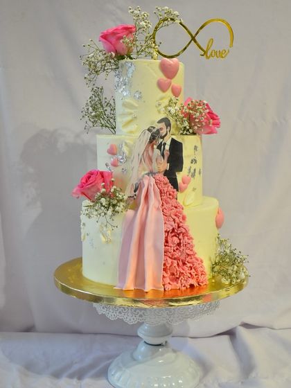 A three-tier engagement cake designed to capture a love story. This custom creation features an edible print of the couple, a flowing ruffled gown detail made of fondant, fresh pink roses, and an infinity love topper.