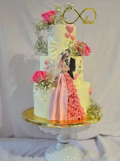 A three-tier engagement cake designed to capture a love story. This custom creation features an edible print of the couple, a flowing ruffled gown detail made of fondant, fresh pink roses, and an infinity love topper.