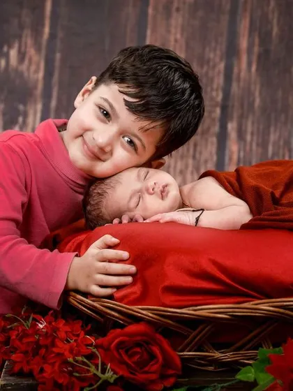 A big brother proudly watches over his sleeping newborn sibling. The rich red tones and rustic wooden background create a warm and loving atmosphere.