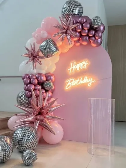 A disco party theme with a pink backdrop, neon sign, and balloon arch featuring silver disco balls and starburst balloons.