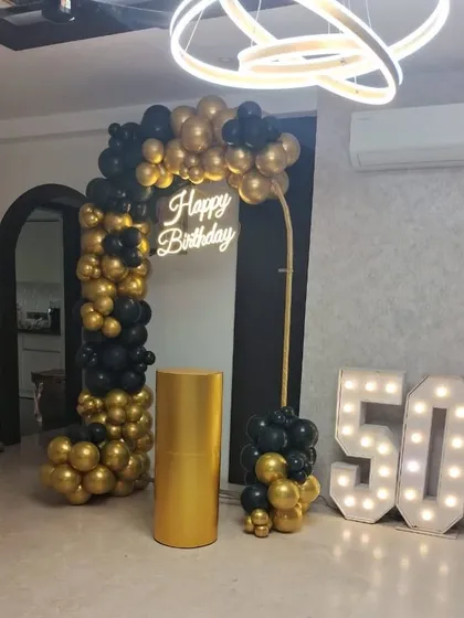 An elegant and modern 50th birthday party decoration. The setup includes a black and gold chrome balloon arch, a 'Happy Birthday' neon sign, and large light-up marquee numbers to mark the milestone.