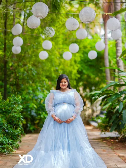 A dreamy solo portrait of the mother-to-be in a light blue gown, standing in a garden path with hanging lanterns.