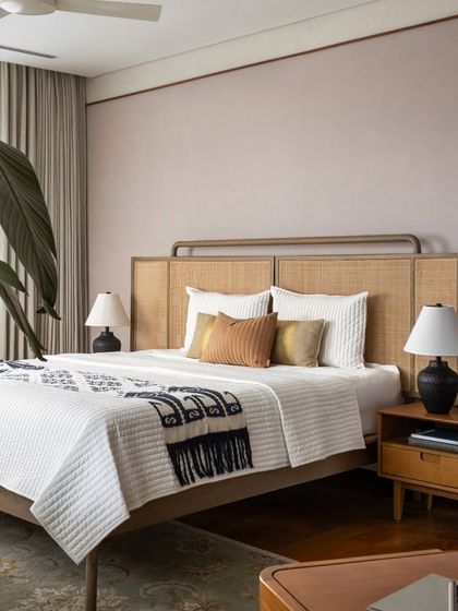 This bedroom balances a contemporary rattan headboard with a soft, dusty pink wall. The space feels refined and inviting, with a mix of textures from the quilted bedspread to the woven rug.