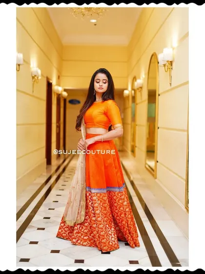 A beautiful shot of Chaitra Vasudevan in a custom orange lehenga, showcasing the outfit's elegant silhouette in a grand hallway. This is a perfect example of festive wear that is both traditional and modern.