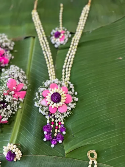 A close-up of a vibrant pink and purple floral necklace and maang tikka. The design uses a large central flower motif surrounded by baby's breath for a full and festive look.