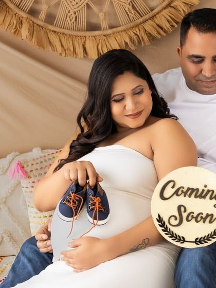 A cozy indoor boho setup. The couple sits together, holding a "Coming Soon" sign and a tiny pair of baby shoes, announcing their pregnancy in style.