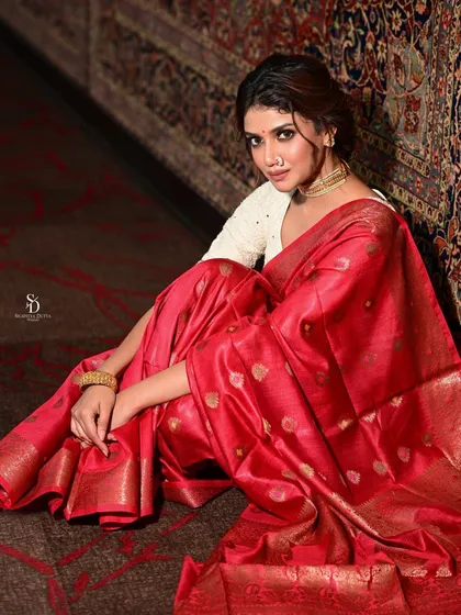 A full-length seated portrait, showing the beautiful drape of the red silk saree. The lighting highlights the sheen of the fabric and its intricate gold motifs.