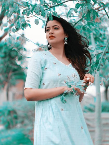 A bright and airy outdoor portrait with a cool, teal-toned color palette. The model stands among light green leaves, creating a fresh and serene atmosphere that complements her light blue dress and gentle expression.