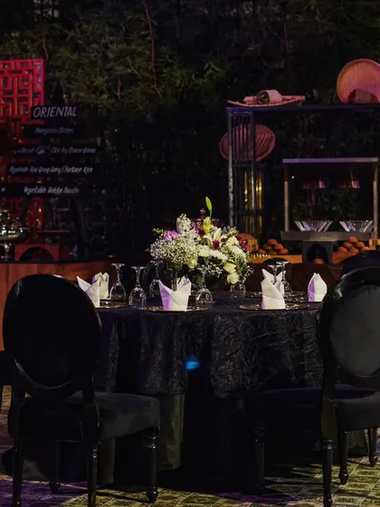 An elegant dining setup with black velvet chairs and tablecloths, creating a sophisticated canvas for the floral centerpieces and the elaborate food stations in the background.