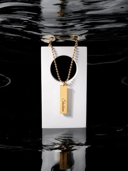 An artistic shot of a gold bar pendant with a script engraving, showcasing its waterproof quality. The reflection in the water emphasizes the pendant's glossy, durable finish.