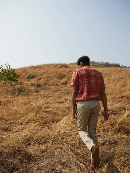 A menswear campaign shot for Burnt Umber Fashion. The model walks through a dry, golden field, creating a rustic and masculine image that perfectly complements the casual, earthy tones of the plaid shirt.