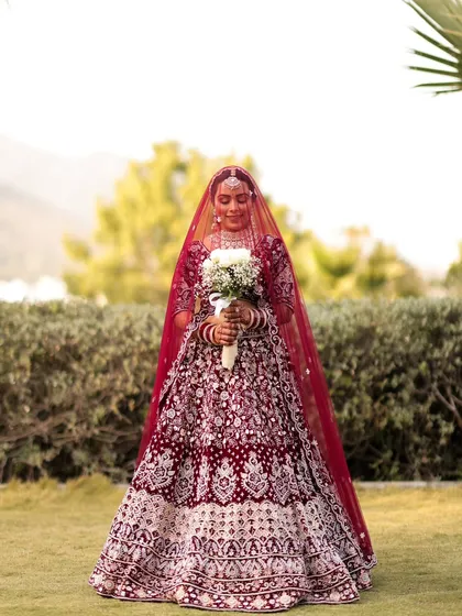 A full-length portrait of the bride in her intricately embroidered maroon lehenga, holding a white bouquet. The outdoor garden setting provides a soft, natural backdrop.