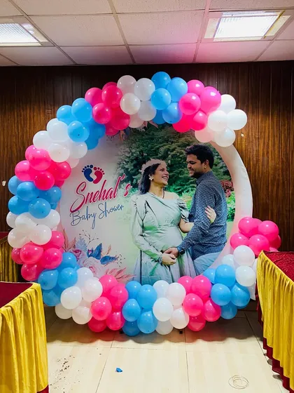 A personalized photo booth for a baby shower, featuring a circular backdrop with the couple's maternity photo, framed by a pink, blue, and white balloon garland.