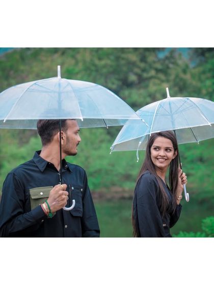 A playful pre-wedding shot of a couple with transparent umbrellas, sharing a glance against a backdrop of lush greenery.