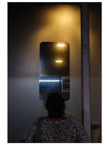 A woman's silhouette is seen as she looks into a small window on a door, with lights reflected in the glass. The image is mysterious and plays with reflections and shadows.