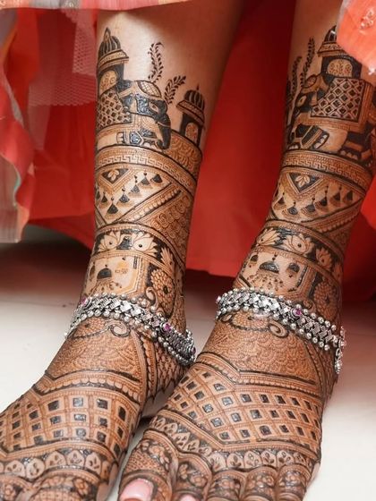 A close-up of intricate bridal mehandi on the feet, adorned with silver anklets. The design features baraat scenes and detailed jaali patterns.