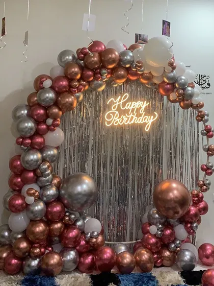 This is how we bring the party to you. We created this luxe balloon arch for a special birthday celebration at home, using a mix of metallic rose gold, silver, and white balloons, complete with a 'Happy Birthday' neon sign against a silver fringe curtain.