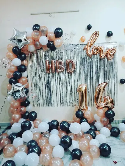 A trendy 14th birthday or anniversary setup. This decoration uses a silver fringe backdrop, rose gold foil letters and numbers, and a stylish black, white, and rose gold balloon arrangement.