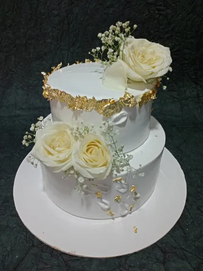 An elegant two-tiered cake adorned with fresh white roses and delicate baby's breath. The torn-edge gold leaf detailing adds a touch of rustic glamour, ideal for weddings or anniversary celebrations.