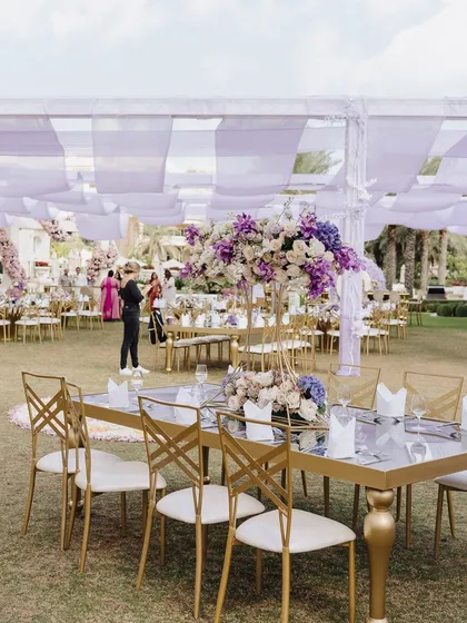 An elegant outdoor dining setup with gold and white chairs, mirrored tables, and tall purple and white floral centerpieces.
