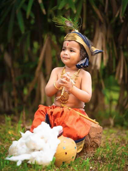 A playful little Krishna enjoying his butter in an outdoor setting. The natural greenery provides a beautiful backdrop that evokes the fields of Vrindavan.