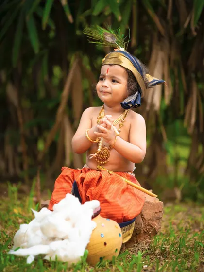 A playful little Krishna enjoying his butter in an outdoor setting. The natural greenery provides a beautiful backdrop that evokes the fields of Vrindavan.