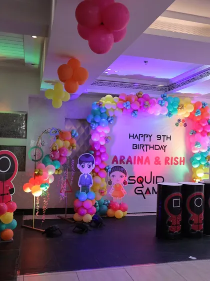 The main stage area for the 'Squid Game' themed party. The setup includes a colorful balloon arch, character cutouts, and themed pedestals, creating a fun and immersive environment.
