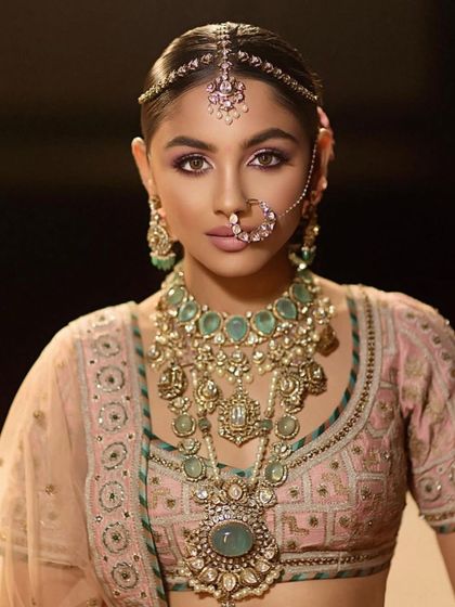 A close-up of a soft and romantic bridal look. The makeup features a subtle purple smokey eye and glowing skin, perfectly paired with a pastel pink lehenga and mint green jewellery.