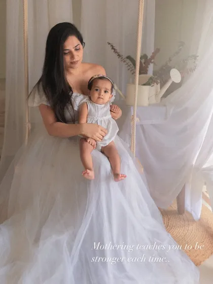 A mother holds her baby, both styled in beautiful white outfits from my client closet. The sheer curtains and natural elements create a serene and timeless atmosphere.