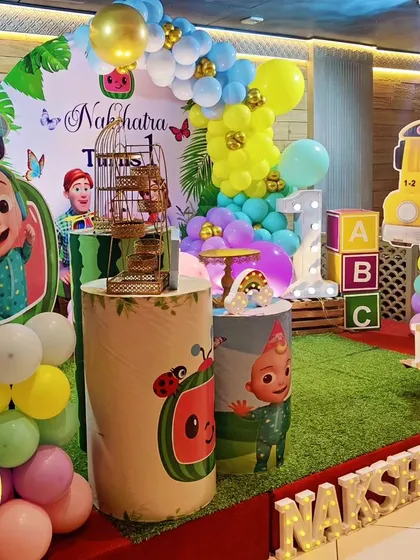 A full stage setup for a Cocomelon-themed first birthday. The decor features a faux grass floor, the iconic yellow bus, and a 'NAKSHATARA' marquee.