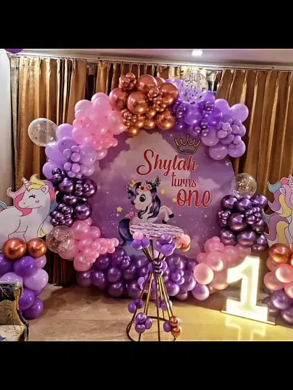 A magical unicorn-themed party perfect for a first birthday. This decoration features a custom backdrop with a beautiful unicorn illustration, a balloon arch in shades of purple, pink, and rose gold, and a light-up number '1' to mark the special milestone.