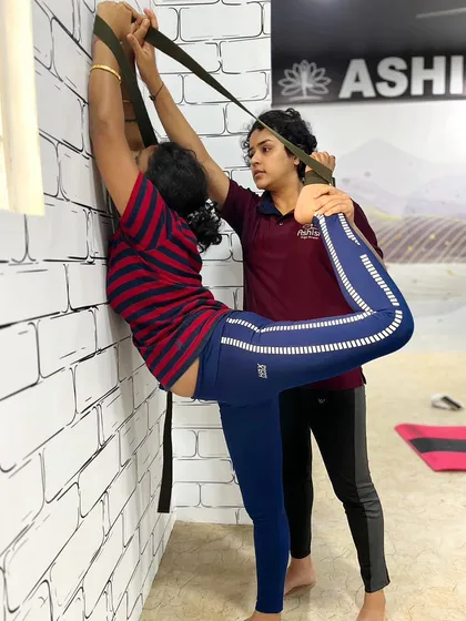 An instructor uses a wall and straps to help a student with a deep standing backbend. This technique provides stability and allows for a gradual, safe increase in flexibility.