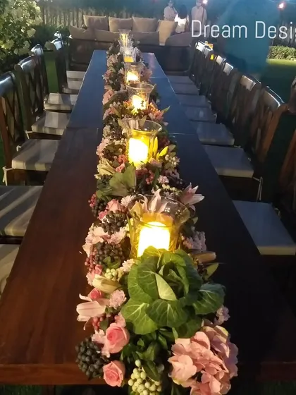 A long wooden table set for an outdoor dinner. The centerpiece is a continuous runner of lush flowers, greenery, and candles in hurricane lamps, creating a rustic yet romantic look.
