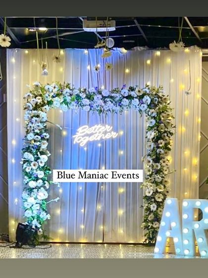 A modern and chic engagement backdrop with a square floral frame of white flowers. The 'Better Together' neon sign, marquee letters, and hanging lights create a fun and stylish photo opportunity.