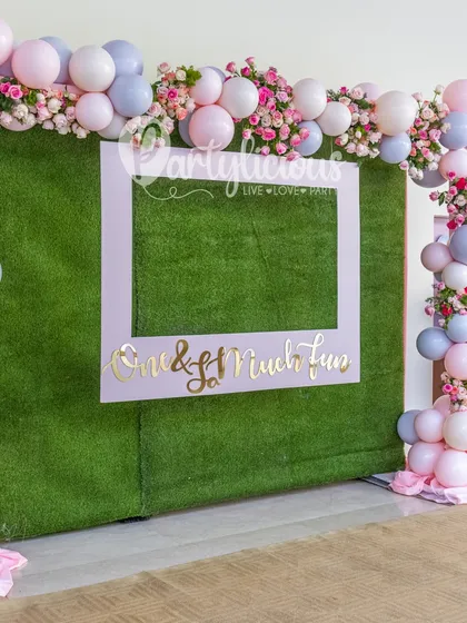 A fun and interactive photo booth. We created this custom frame, "One & So Much Fun," set against a grass wall and framed with a pink and lilac floral balloon garland.