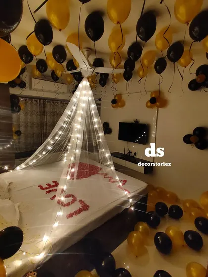 For this romantic room decoration, we installed a delicate canopy draped with fairy lights over the bed. The look was completed with black and gold balloons floating on the ceiling and scattered on the floor, and a heart shape made from real rose petals on the bed.