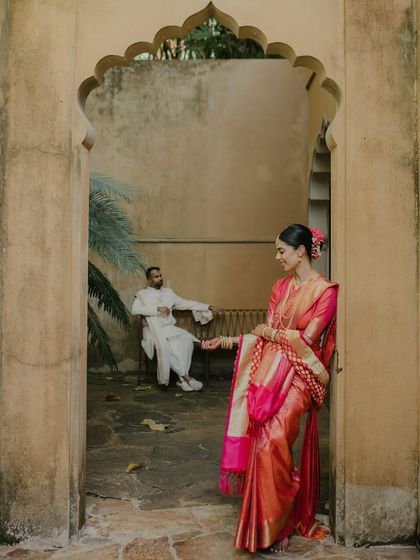 An artistic shot from a Sri Lankan wedding. The bride, in her vibrant Kanjeevaram sari, stands in an archway while the groom waits in the background, creating a sense of anticipation and beautiful composition.