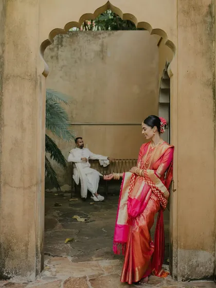 An artistic shot from a Sri Lankan wedding. The bride, in her vibrant Kanjeevaram sari, stands in an archway while the groom waits in the background, creating a sense of anticipation and beautiful composition.
