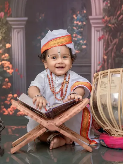 A joyful baby Varkari, happily sitting with his holy book and tabla. The entire setup, with its floral background and traditional props, comes together to create a perfect memory.